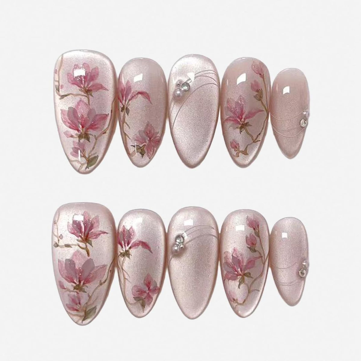 A set of ten almond-shaped press-on nails with a shimmering pale pink base. The design features hand-painted pink magnolia flowers on brown branches, accented with thin silver lines and small white pe