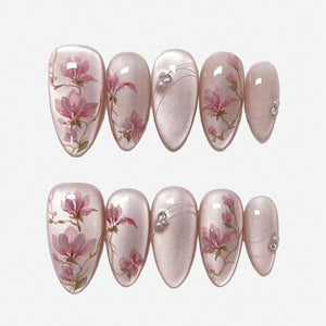 A set of ten almond-shaped press-on nails with a shimmering pale pink base. The design features hand-painted pink magnolia flowers on brown branches, accented with thin silver lines and small white pe