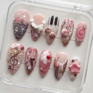 A set of ten press-on nails in a clear plastic case, featuring various pink, brown, and white designs including piano keys, a treble clef, a cherry, stars, hearts, and 3D embellishments like small flowers, bows, and a pink star.