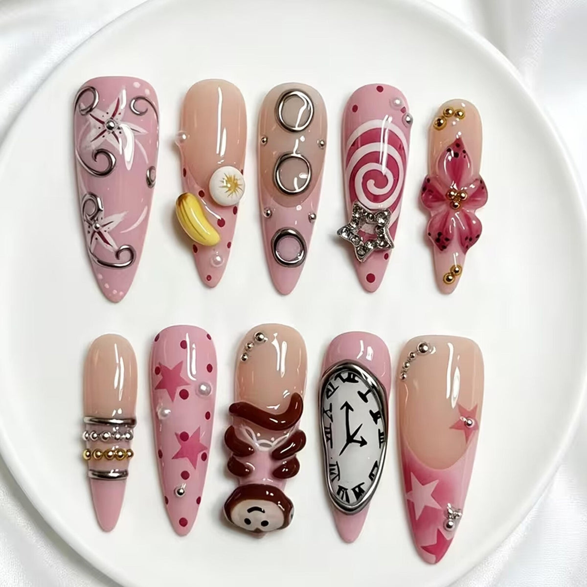 A set of ten almond-shaped press-on nails featuring a pink and nude color palette. Designs include silver swirls, a yellow banana charm, metallic rings, a star charm, a 3D flower, silver beads, pink stars, a monkey figure, and a clock.