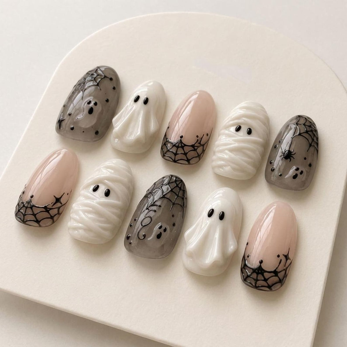 A set of ten press-on nails featuring Halloween designs including white ghosts, wrapped mummies, and sheer grey nails with black spiderweb patterns and small spiders. The nails have a glossy finish and oval shape.