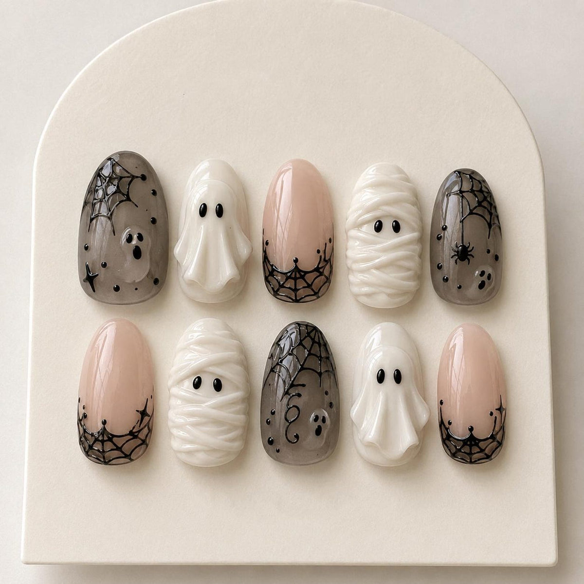 A set of ten press-on nails featuring Halloween designs. The collection includes translucent gray nails with black spiderwebs and ghosts, white nails with raised ghost or mummy textures, and nude nails with black spiderweb tips and star accents.