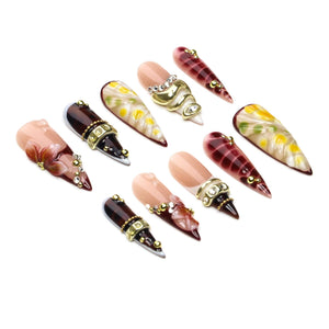 A set of ten stiletto-shaped press-on nails featuring a variety of designs including nude bases with hand-painted floral art, deep burgundy tones with gold metallic bands, and clear rhinestones, all arranged on a plain white background.