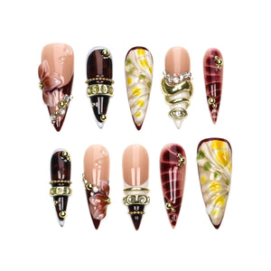 A set of ten stiletto-shaped press-on nails featuring a variety of designs including nude bases with 3D floral accents, dark brown tortoiseshell patterns with gold metal bands, white and yellow floral motifs, and deep burgundy swirls.