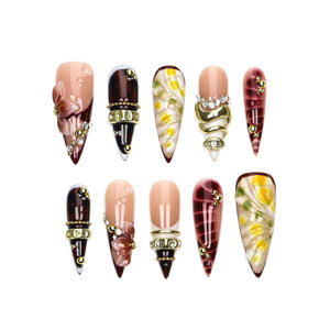 A set of ten stiletto-shaped press-on nails featuring diverse designs including dark brown tortoiseshell patterns, nude bases with 3D floral accents, gold metallic bands, clear rhinestones, and white petals with yellow centers.