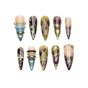 A set of ten press-on nails featuring pointed tips, decorated with a mix of deep purple, gold, and blue tones. The designs include intricate metallic filigree, embedded rhinestones, cross motifs, and layered ring-like embellishments.