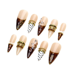 A set of ten stiletto-shaped press-on nails featuring a nude base with brown tortoise shell patterns, black polka dots, and hand-painted floral accents. The designs are embellished with gold-toned metallic chains and small rhinestones.
