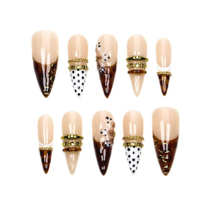 A set of ten stiletto-shaped press-on nails featuring a nude base with various designs including dark brown tortoise shell patterns, black polka dots on white, gold-toned metallic bands, and three-dimensional floral accents.