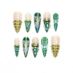 A set of ten press-on nails featuring a variety of designs in shades of green, cream, and gold. The nails include pointed and rounded shapes adorned with metallic gold studs, green gemstone accents, intricate gold filigree, and patterns.
