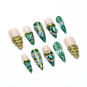 A set of ten press-on nails featuring a nude base and a pointed green tip with a crocodile skin pattern. The nails are decorated with gold metal accents, small clear rhinestones, and green heart-shaped or star-shaped gems.