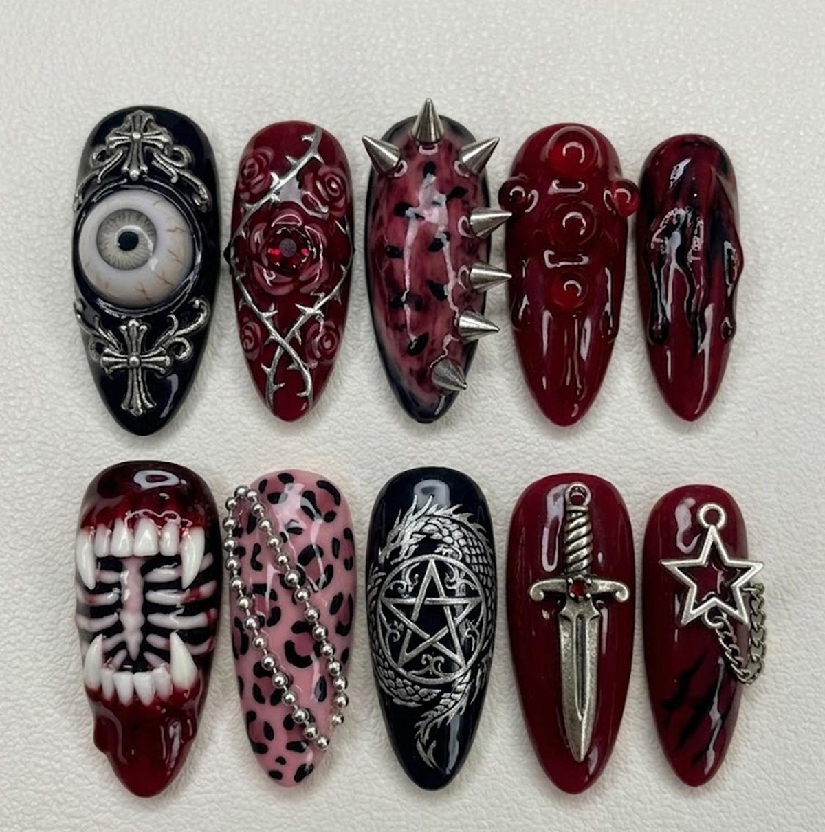A set of ten press-on nails featuring dark red and black designs. Elements include a realistic eyeball, silver crosses, roses with thorns, metal spikes, blood-like drips, white teeth, a skeleton ribcage, pink leopard print, a dragon pentagram, a silv
