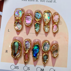 A set of ten press-on nails with a light pink base, each featuring irregular, iridescent abalone shell fragments encased in raised, textured gold-toned metal borders. The nails are arranged in two rows on a display card labeled L and R.