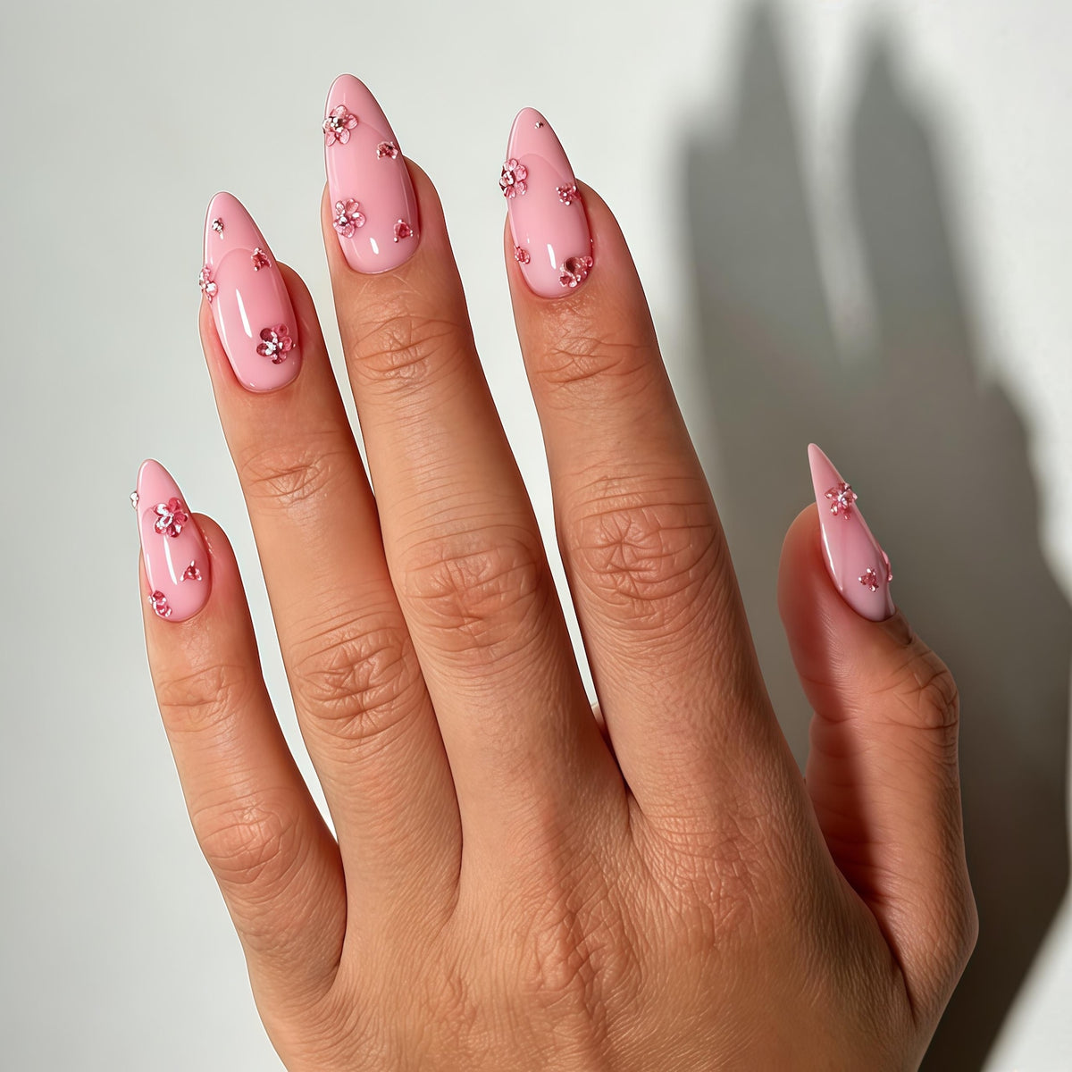 Almond-shaped nails with a glossy light pink base coat, each featuring small, three-dimensional pink flower embellishments and tiny clear rhinestones scattered across the surface of the nail beds for a textured, decorative finish.
