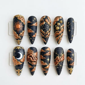A set of ten almond-shaped press-on nails featuring a black base with a celestial theme. The designs include raised copper-colored clouds, a sun, a crescent moon, constellations, and gold-painted stars against a speckled night sky.