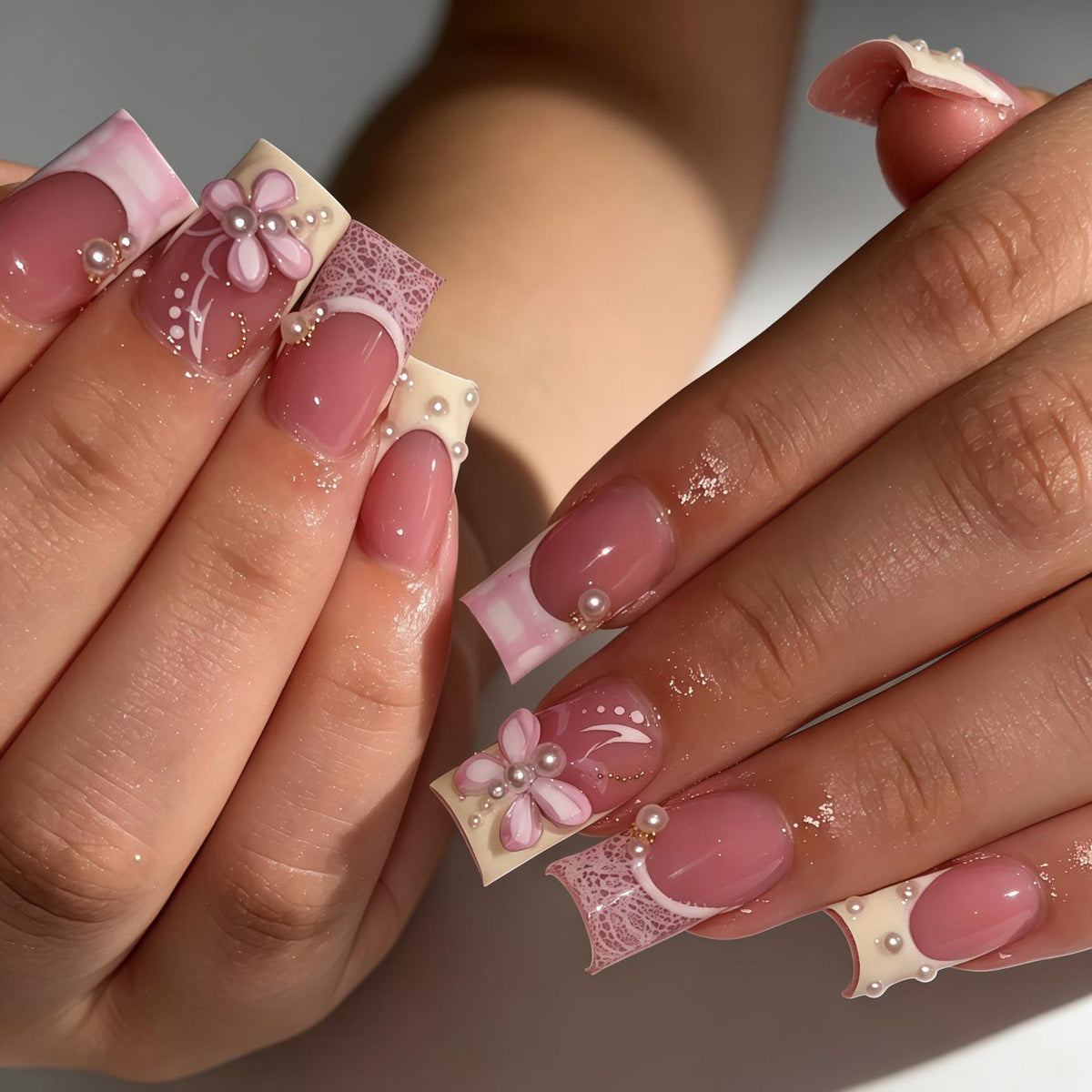 Square-shaped artificial nails featuring a pink base with white French tips. The design includes intricate white lace patterns, three-dimensional pink flower appliqués, and small white pearl accents placed along the nail beds and tips.