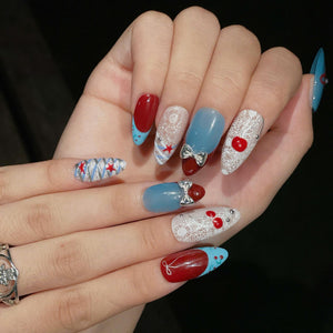 Almond-shaped nails featuring a mix of red and blue polish, white lace patterns, and silver bow charms. Some nails display red star accents, while others feature small red cherry motifs and silver studs, creating a detailed manicure.
