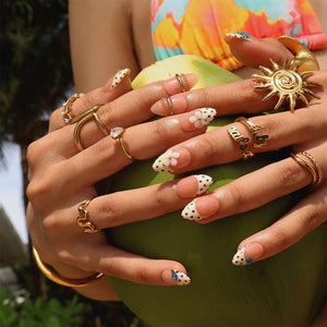 A collection of gold-toned rings worn on fingers with almond-shaped nails featuring a French manicure style with white tips, blue polka dots, and small white floral accents. The rings include a sun-shaped design and script text.