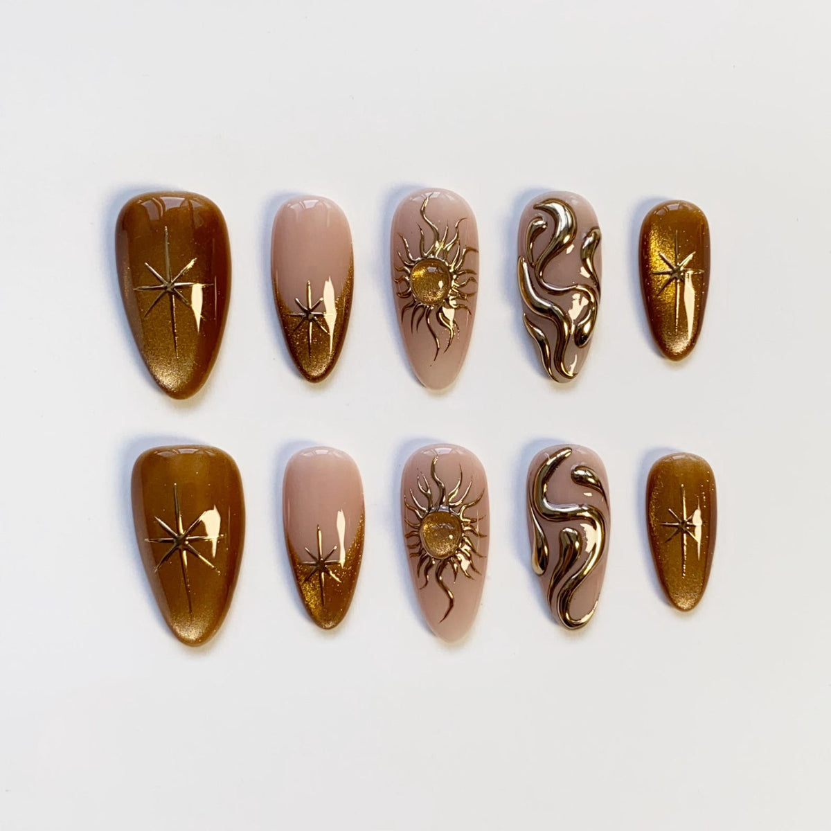A set of ten artificial nails in various designs. Some nails are a metallic gold color with starburst accents. Others are a nude pink with gold sun and swirl designs. The nails are oval-shaped.