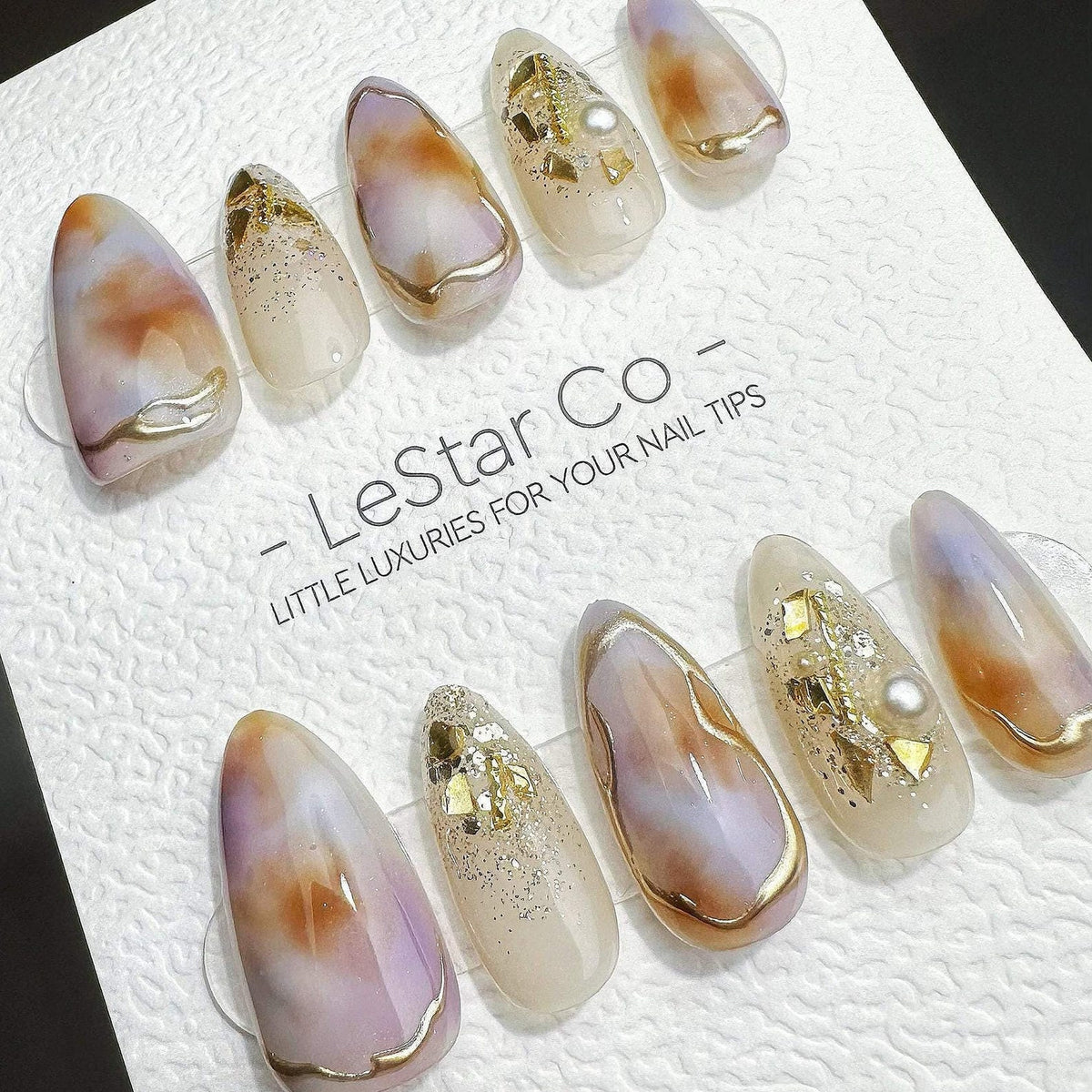 A set of ten almond-shaped press-on nails featuring a marbled design in shades of purple, brown, and cream, accented with gold foil flakes, glitter, and small white pearls. The nails are displayed on 