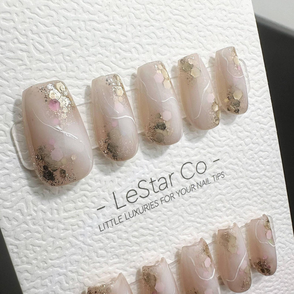 A set of press-on nails featuring a translucent nude base with gold and pink hexagonal glitter accents, white swirl line details, and a glossy finish. The nails are displayed on a white card labeled L