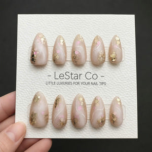 A set of ten almond-shaped press-on nails in a soft nude base color, featuring gold and pink hexagonal glitter accents and thin white swirl lines. The nails are displayed on a white textured card labe