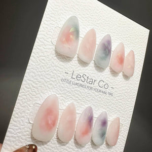 A set of ten almond-shaped press-on nails with a translucent, milky base featuring soft watercolor-style swirls of pink, purple, and green. The nails are displayed on a white textured card labeled LeS