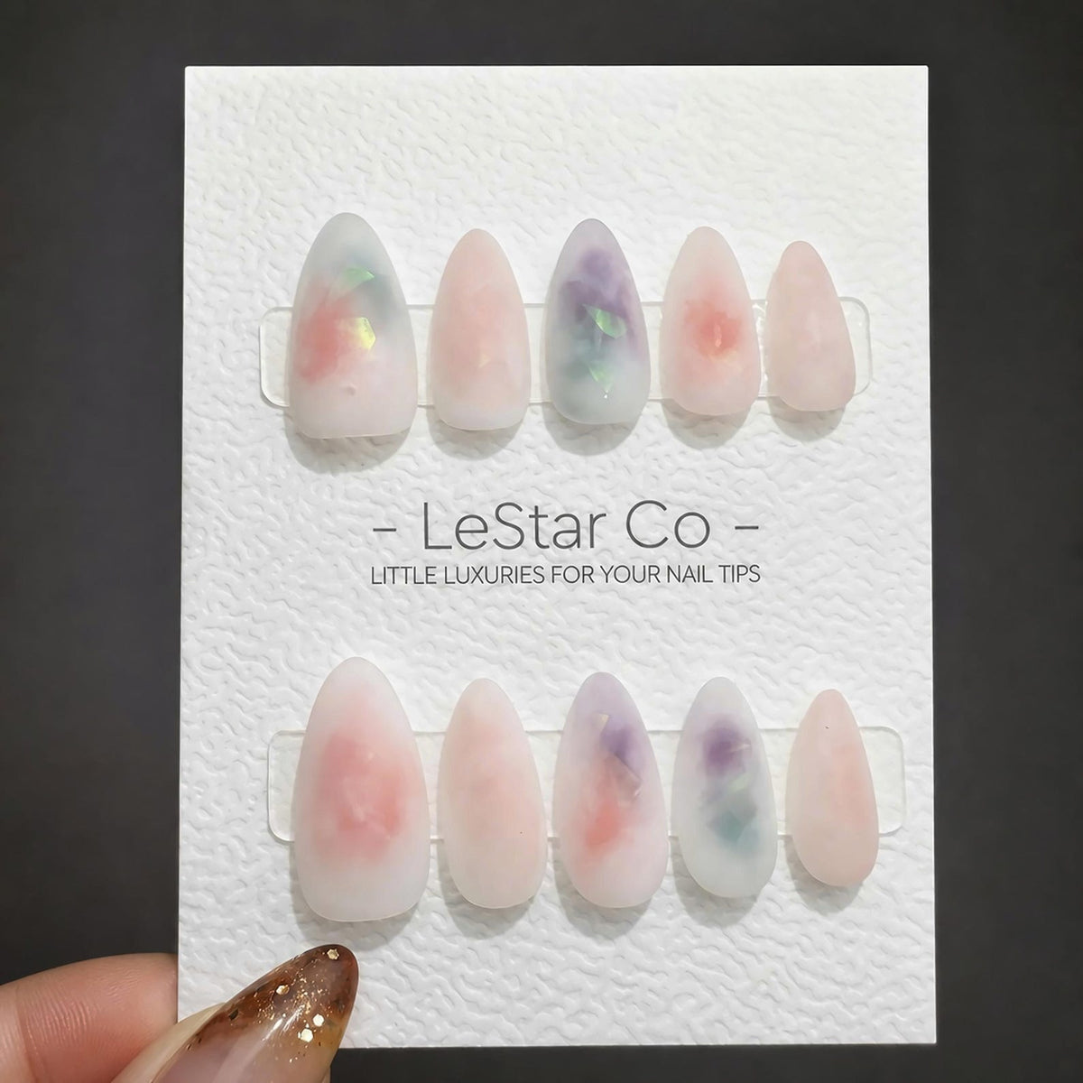 Ten almond-shaped press-on nails with a milky translucent finish, featuring embedded iridescent flakes in shades of pink, purple, and green. The nails are displayed on a white card labeled LeStar Co -