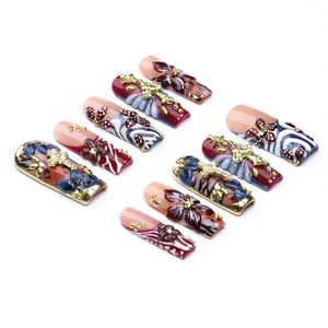 A set of ten press-on nails featuring a variety of designs including deep red, blue, and nude bases with white swirling patterns, dark floral accents, and gold-toned cross embellishments adorned with small clear rhinestones.