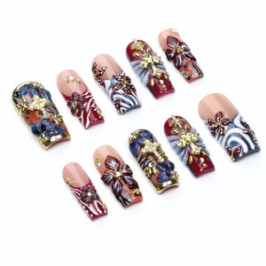 A set of ten press-on nails featuring a nude base with intricate designs including deep red and blue floral patterns, white swirling accents, gold-toned cross embellishments, and small clear rhinestones applied to the surface.