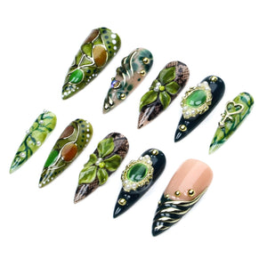 A set of ten stiletto-shaped press-on nails featuring various designs in shades of green, black, and nude. The nails are decorated with 3D floral accents, gold metallic details, faux pearls, intricate lace patterns, and green cabochon gems.