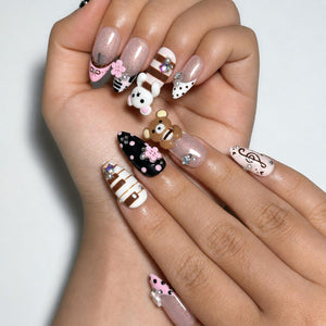 Ten pink, black, brown and white almond press on nails with 3D teddy bear charms, flowers, music notes, polka dots, stripes, pearls and rhinestones on a white background.