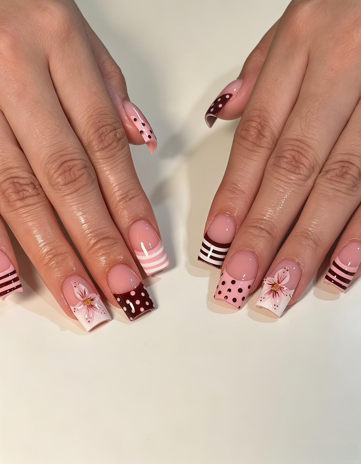 Square-shaped fingernails featuring a French manicure style with a mix of designs. Some nails have dark maroon tips with white polka dots, others have light pink stripes, and some feature a hand-painted white flower with pink accents.