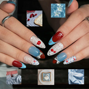 Almond-shaped artificial nails featuring a mix of deep red and light blue polish, white lace patterns, small red cherry accents, and silver-toned bow charms. The nails are displayed with small inset images of cherries, white flowers, striped bedding,