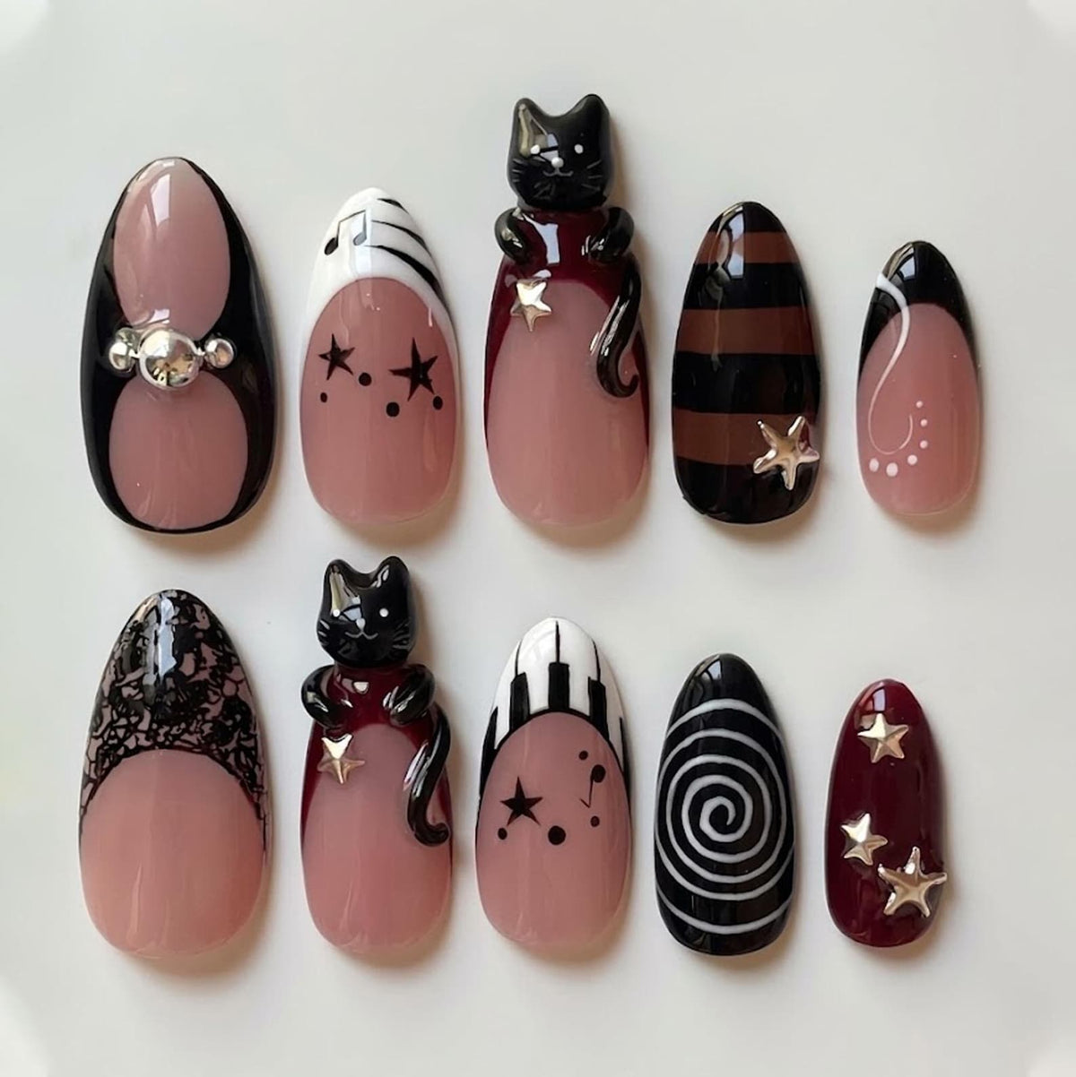 A set of ten press-on nails featuring a nude base with various designs including black cat figures, silver star charms, piano keys, musical notes, black lace patterns, brown and black stripes, and a white spiral on a black background.