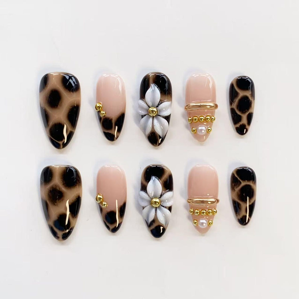 A set of ten press-on nails featuring a mix of designs including brown and black leopard print, solid nude bases with small gold studs, white floral accents with gold centers, and nude nails with horizontal gold bands and pearl beads.