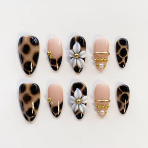 A set of ten press-on nails featuring a mix of designs. Some nails have a brown and black leopard print pattern, while others are a nude base with white 3D flower accents, gold metallic beads, and small white pearls.