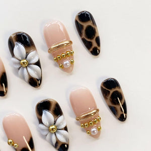 A set of almond-shaped press-on nails featuring a mix of designs. Some nails have a nude base with gold metallic bands, small gold beads, and a single white pearl. Others feature a black and brown leopard print or white floral art.