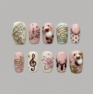 A set of ten press-on nails featuring various designs including a 3D butterfly, musical notes, a 3D cat figure, swirling gold lines, polka dots, a 3D pink flower, and small star accents in shades of cream, soft pink, and brown.