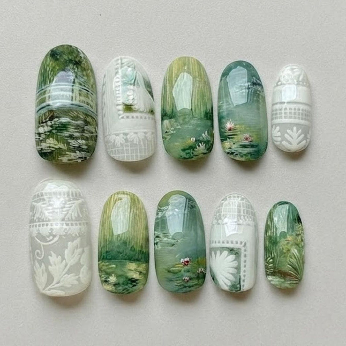 A set of ten press-on nails featuring a variety of designs in shades of green and white. The nails display intricate hand-painted landscapes, water lily motifs, and embossed white lace-like patterns on glossy, oval-shaped surfaces.