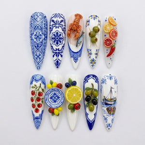A set of ten stiletto-shaped press-on nails featuring Mediterranean-inspired designs. The collection includes blue and white tile patterns, 3D miniature food motifs like lobster, olives, citrus slices, tomatoes, and a sailboat illustration.