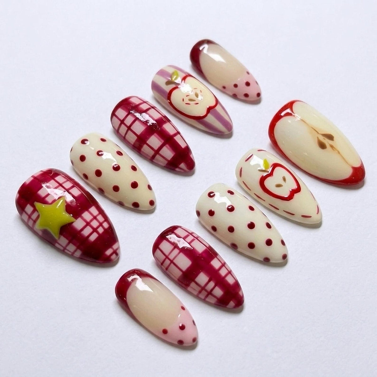 A set of ten almond-shaped press-on nails featuring various designs including red and white plaid patterns, cream-colored surfaces with red polka dots, a yellow star accent, and detailed red apple illustrations on a glossy finish.