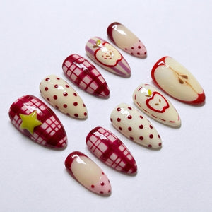 A set of ten almond-shaped press-on nails featuring various designs including red and white plaid patterns, cream-colored surfaces with red polka dots, a yellow star accent, and detailed red apple illustrations on a glossy finish.