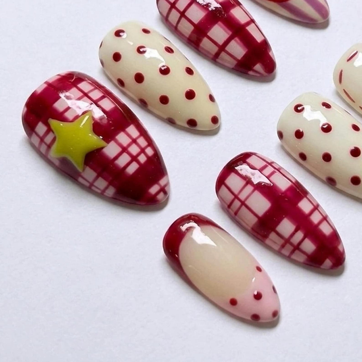 A set of almond-shaped press-on nails featuring a variety of designs including a red and white plaid pattern, cream-colored nails with red polka dots, and a single nail with a yellow star accent over a red plaid background.