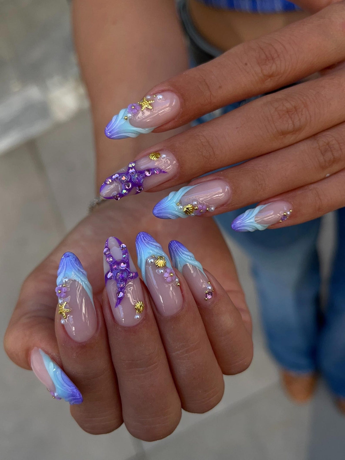 Long almond-shaped nails featuring a nude base with blue and purple gradient tips. The design includes three-dimensional purple starfish, small gold seashell charms, tiny white pearls, and clear rhinestones applied to the nail surface.