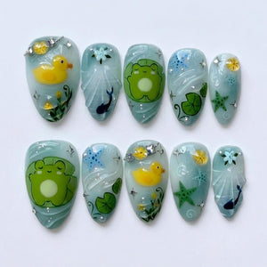 A set of ten press-on nails featuring a light blue base with pond-themed designs including yellow rubber ducks, green frogs on lily pads, blue and green starfish, small dark blue whales, and tiny white floral accents with silver glitter.