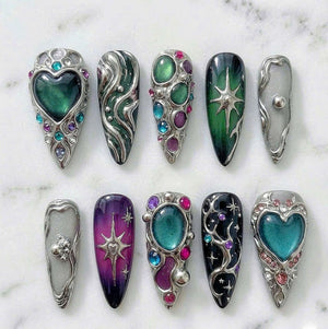 A set of ten press-on nails featuring intricate 3D silver metal accents, embedded colorful gemstones in shades of green, purple, and blue, and glossy finishes. Designs include heart shapes, starbursts, and swirling metallic patterns.