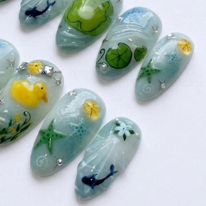 A set of press-on nails featuring a light blue base with various aquatic designs including a yellow rubber duck, green starfish, lily pads, small blue flowers, and dark blue whale silhouettes, accented with tiny silver star-shaped studs.