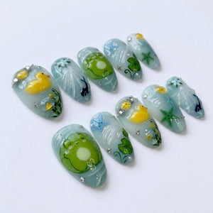 A set of ten almond-shaped press-on nails with a light blue base. The designs feature hand-painted yellow rubber ducks, green frogs on lily pads, blue and green starfish, and small white floral accents, all embellished with tiny silver rhinestones.