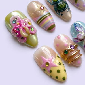 A collection of almond-shaped press-on nails featuring diverse designs including a 3D pink flower with rhinestones, a beige nail with a star and stripes, a green beetle, a polka dot nail with a pink star, and a gold ring-adorned nail.