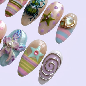 A collection of almond-shaped press-on nails featuring diverse 3D designs including a metallic green beetle, a purple polka-dot nail with a green star, a blue and pink striped nail with a shell accent, a floral motif, and a silver spiral.